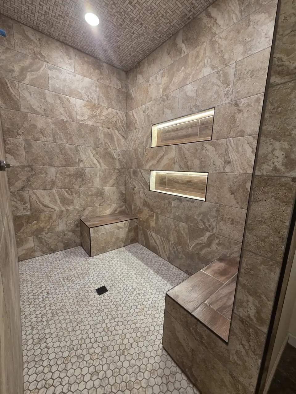 Walk-in tile shower with travertine-look walls, two lit niches, two tiled benches and a hexagon mosaic floor, flooring and tile Greenbrier